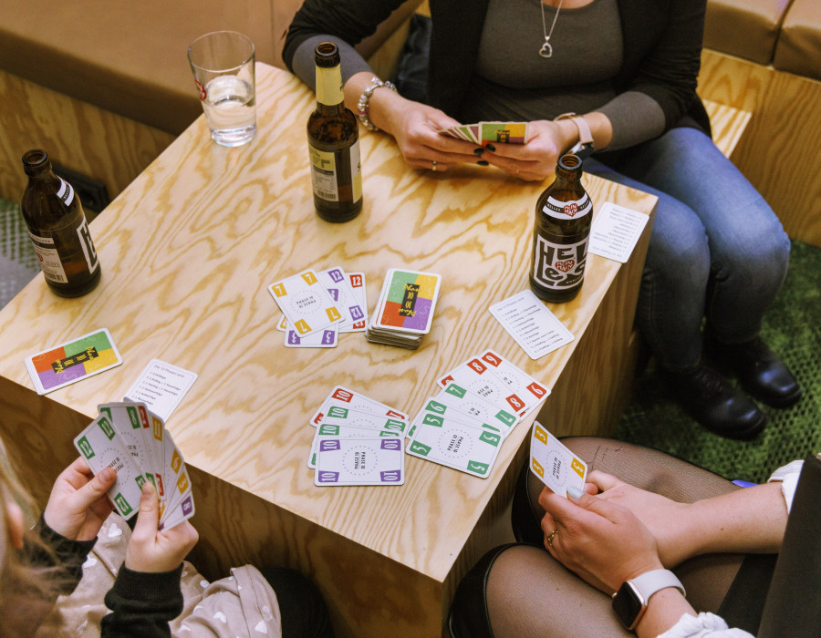 Personen spielen ein Kartenspiel an einem Holztisch mit Bierflaschen, Gläsern und bunten Karten.