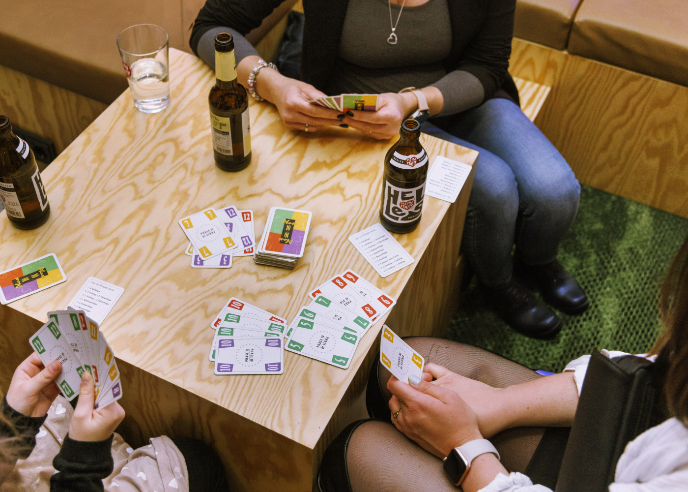 Vier Personen sitzen um einen Holztisch und spielen ein buntes Kartenspiel, dazu Bierflaschen und Gläser.