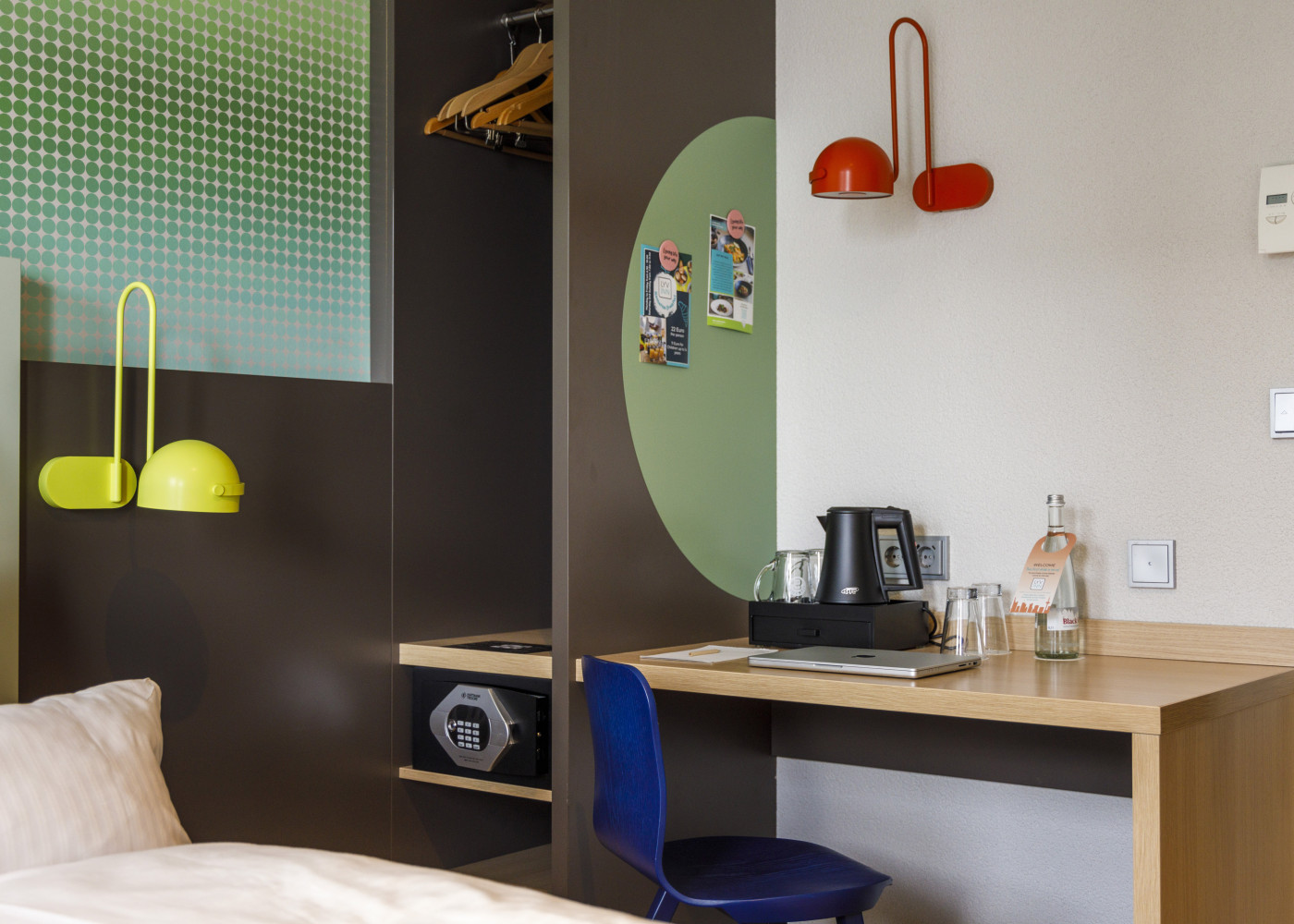 Frisch gemachtes Bett, daneben ein Tisch und eine auffällige gelbe Wandlampe im Hotelzimmer.