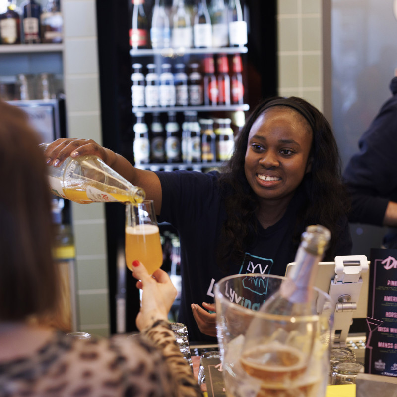 Freundliche Mitarbeiterin schenkt einem Gast ein Glas Saft ein, im Hintergrund ein Getränkekühlschrank