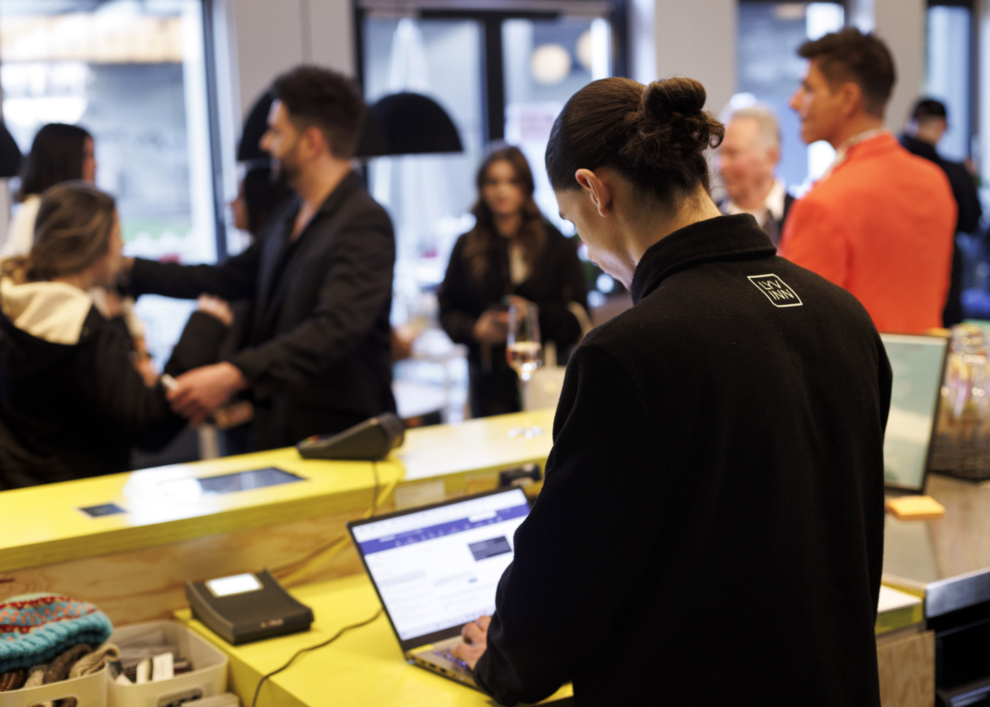 Person arbeitet an einem Laptop am Empfangstresen, während im Hintergrund Gäste lebhaft interagieren.