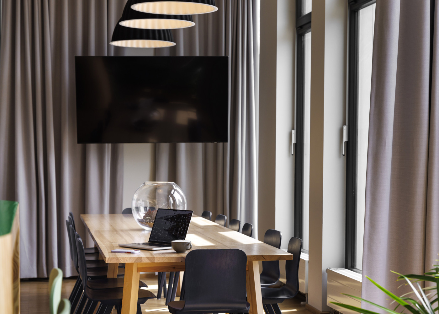 Langer Besprechungstisch mit Laptop, Stühlen und großer Monitorwand, flankiert von Vorhängen und Fenstern.