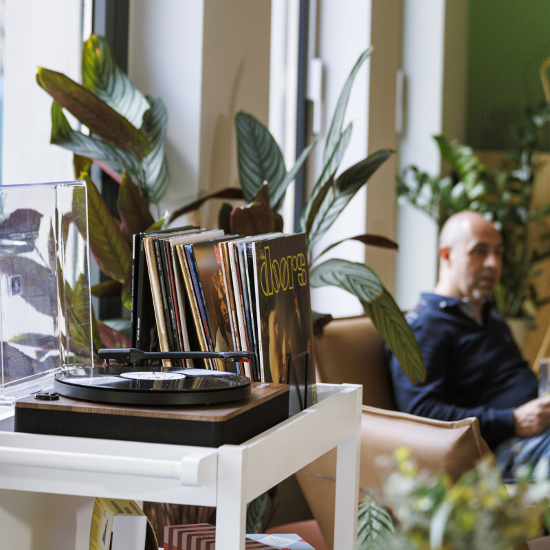 Schallplattenspieler mit Vinyl-Sammlung, umgeben von Pflanzen, während ein Mann entspannt in der Hotellounge sitzt.