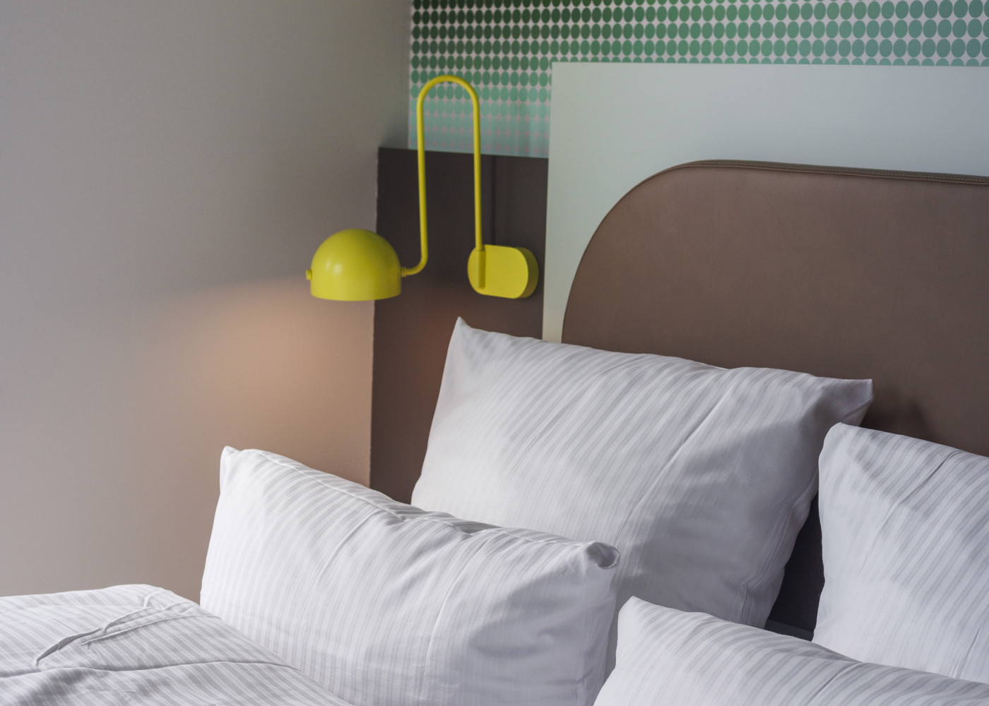 Gemachtes Bett mit weißen Kissen und Decke, daneben eine moderne gelbe Wandlampe im Hotelzimmer.
