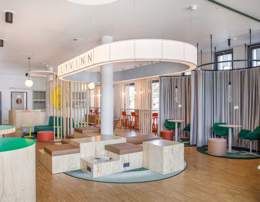 Lichtdurchfluteter Lounge-Bereich mit Holzsitzmöbeln, farbigen Akzenten und großem LYV INN Schild.