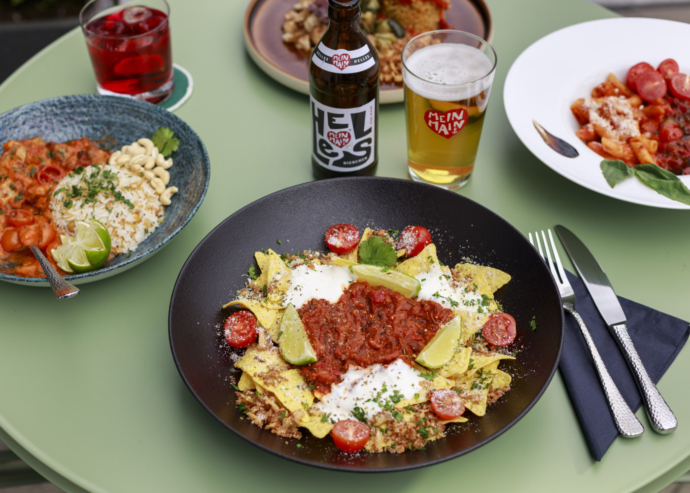 Tisch mit verschiedenen bunten Gerichten, Bier, Getränken und Besteck – angerichtet auf grünem Outdoor-Tisch.