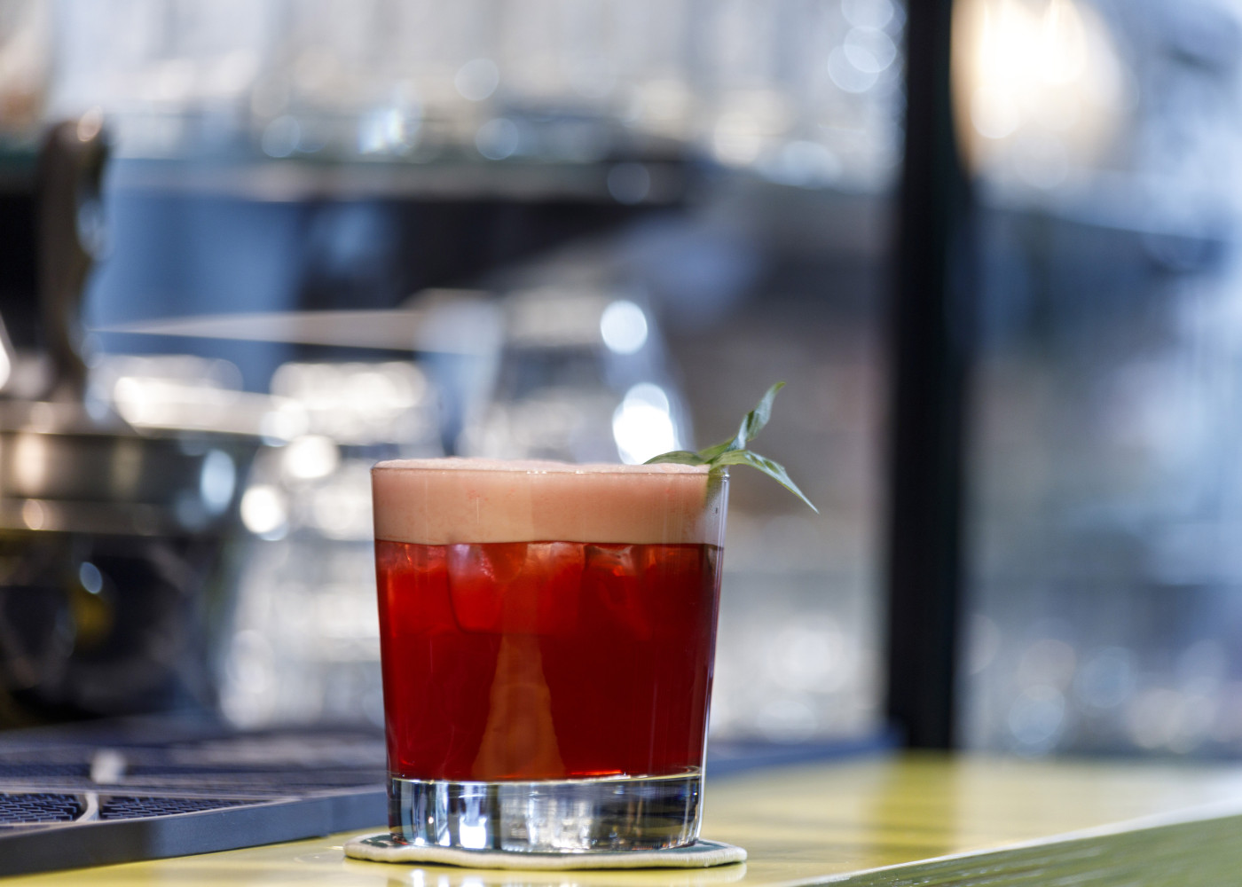 Roter Cocktail mit Schaumkrone und Kräutergarnitur in einem Glas, serviert auf einer Bar mit Tageslicht.