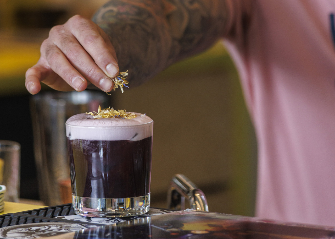Tätowierte Hand garniert einen Cocktail mit violettem Schaum vorsichtig mit getrockneten Blüten