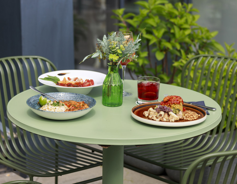 Drei bunte Gerichte und ein rotes Getränk auf grünem Tisch mit Blumen, umgeben von Gartenstühlen.