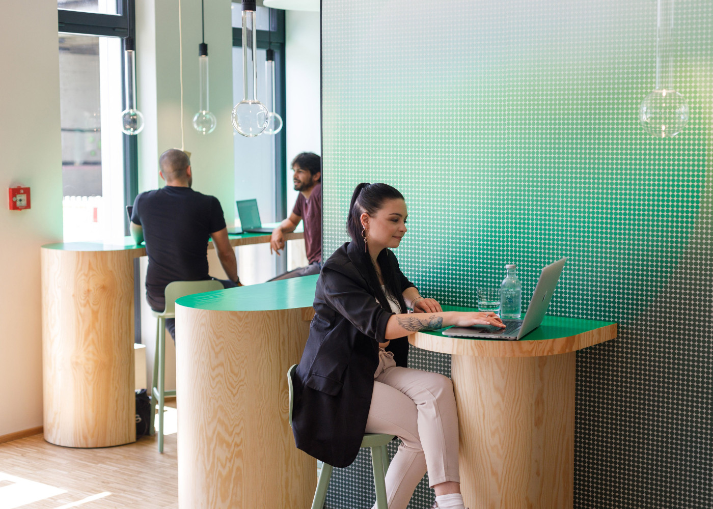 Menschen arbeiten an Laptops auf hohen Holztresen in einem modernen Raum mit grün strukturierter Wand.