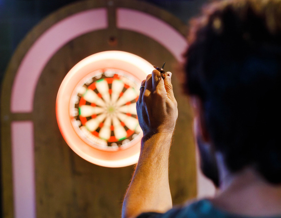 Mann steht vor einer beleuchteten elektronischen Dartscheibe und wirft einen Dartpfeil im Spielbereich.