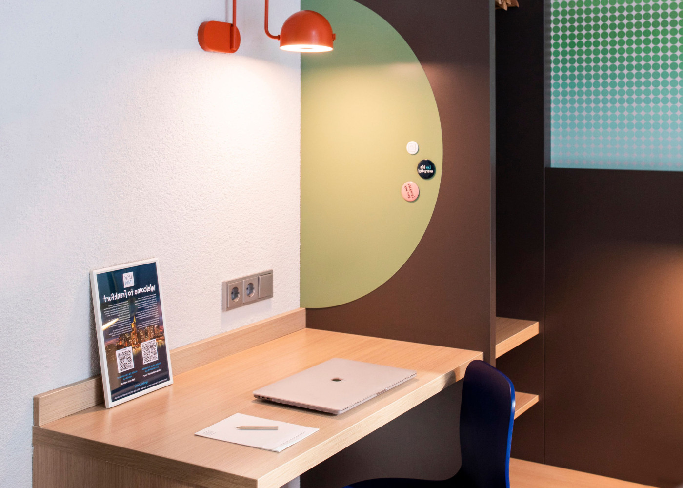 Aufgeräumter Holzschreibtisch mit Laptop, Block, oranger Lampe und Prospekt vor farbiger Wandgestaltung.