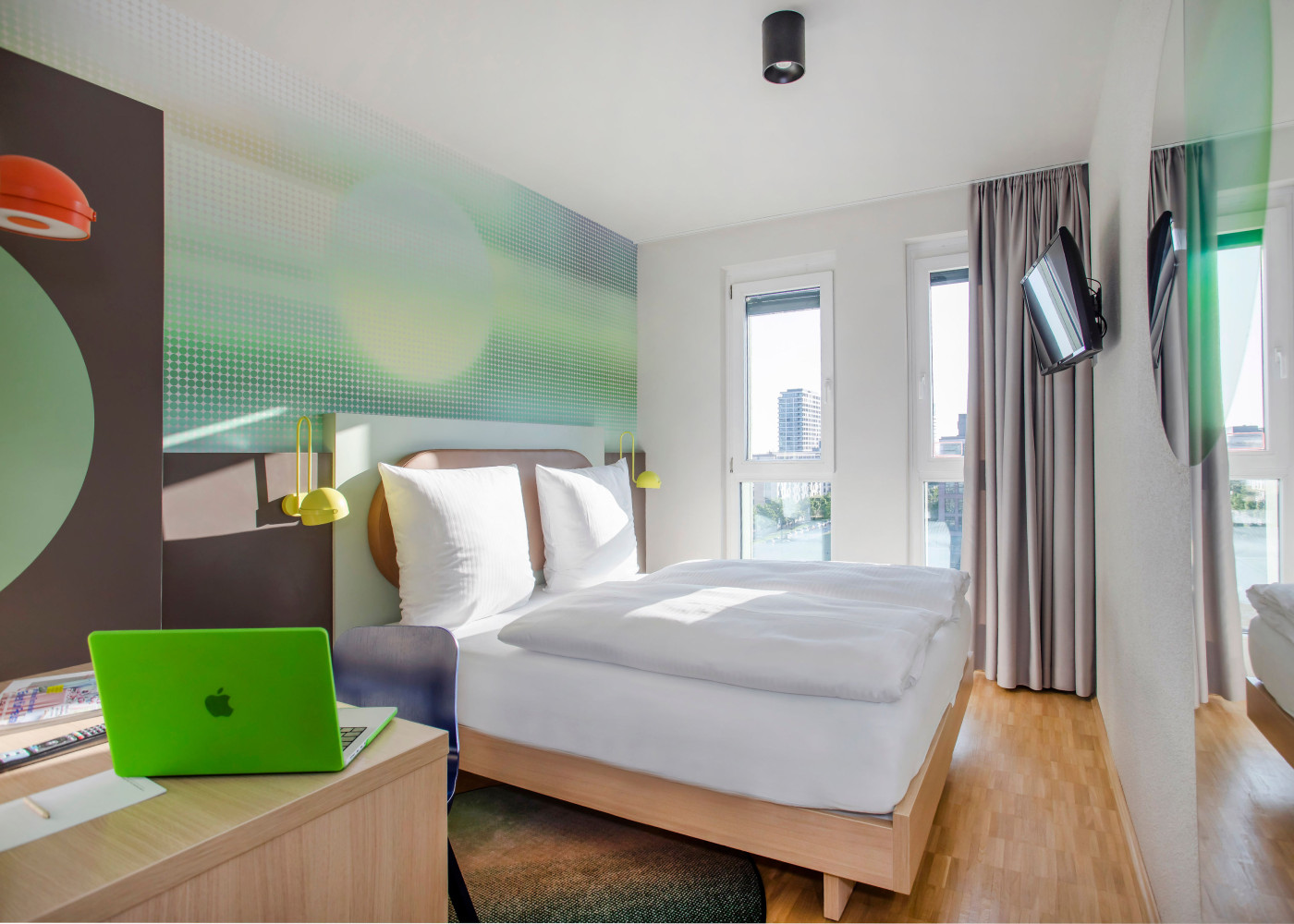 Helles Hotelzimmer mit großem Bett, Fensterfront, grünem Laptop und bunter Wandgestaltung mit Lichtakzenten.