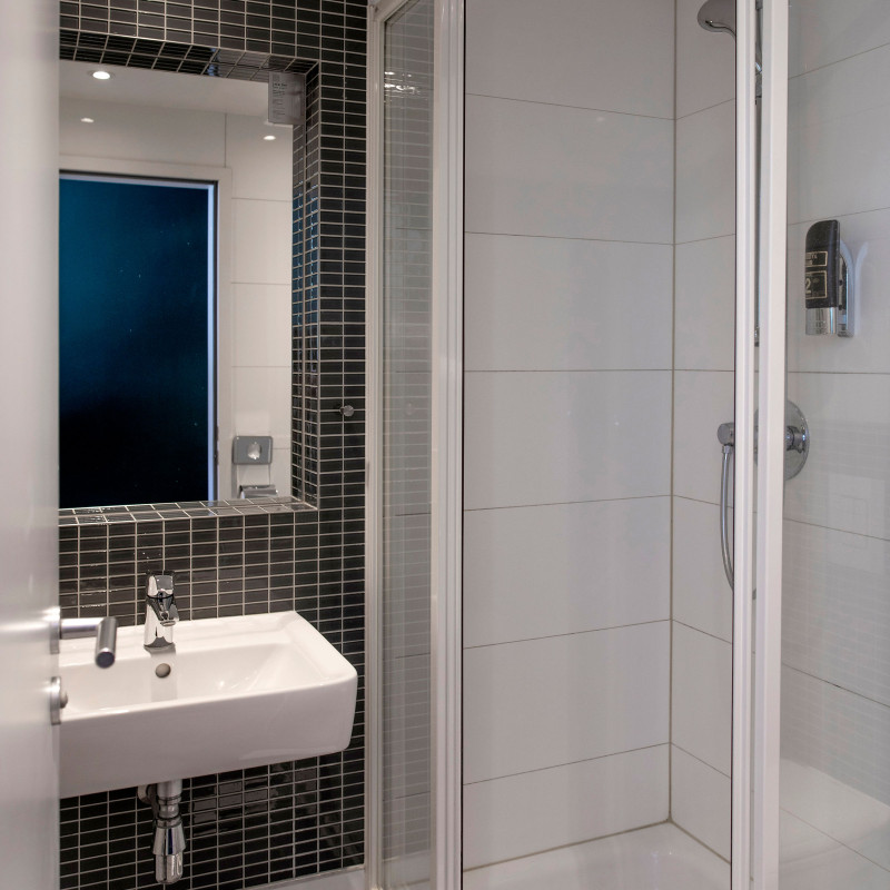 Weiß gefliestes Hotelbad mit großer Glasdusche, Waschbecken und dunkler gefliester Wand hinter dem Spiegel.