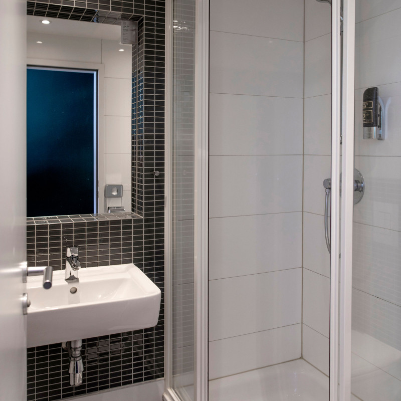 Weiß gefliestes Bad mit Glasdusche, Spiegel und Waschbecken, schwarzer Fliesenakzent hinter dem Spiegel.