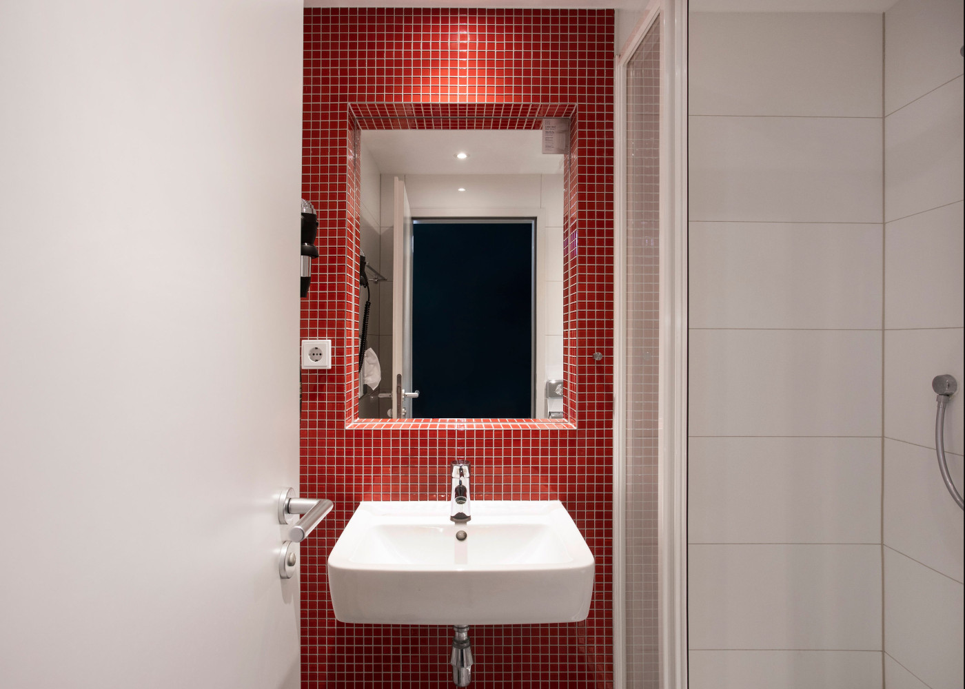 Kleines Badezimmer mit weißem Waschbecken und Spiegel vor einer roten Fliesenwand und Duschabtrennung.