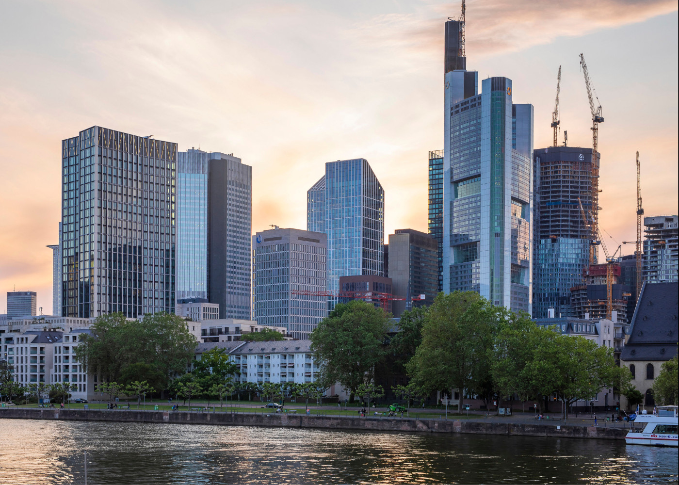 Skyline moderner Hochhäuser in Frankfurt bei Sonnenuntergang, gesehen vom Flussufer mit Bäumen davor.