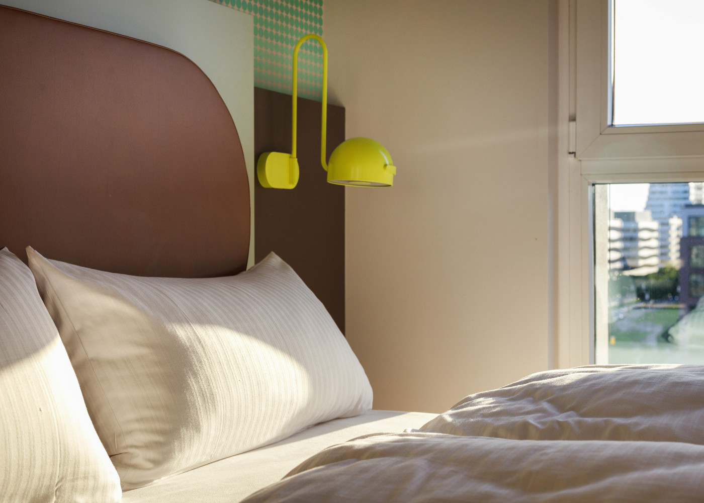 Weiches Bett mit weißer Bettwäsche und gelber Wandlampe, daneben Fenster mit Ausblick auf die Stadt.