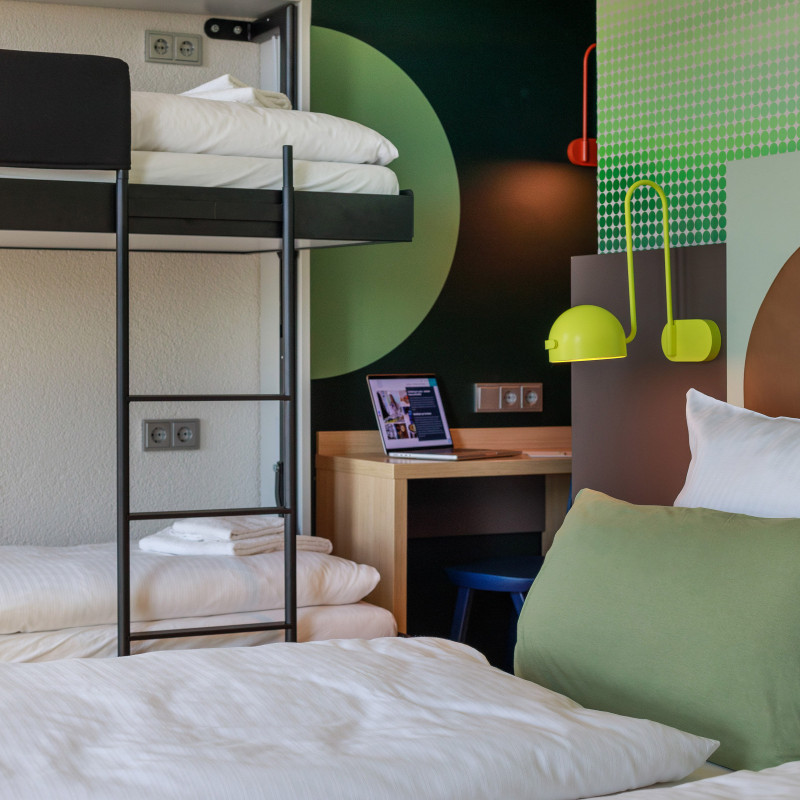Hotelzimmer mit Doppelbett, Etagenbett, grünem Wanddesign und Schreibtisch mit Laptop im Hintergrund.