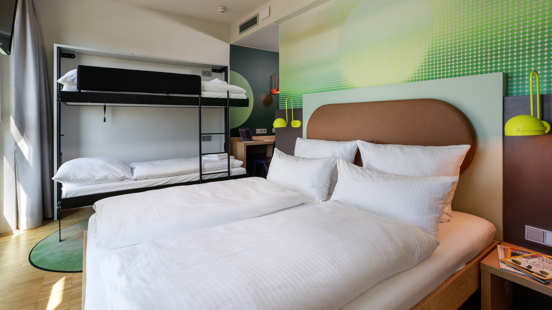 Blick auf ein Doppelbett vor einer grünen Wand und ein Stockbett im Hintergrund.