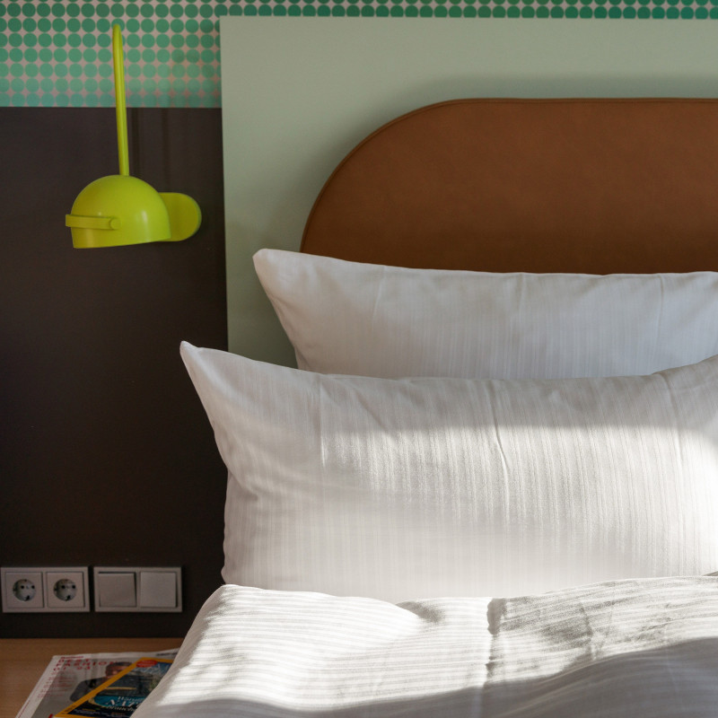 Nahaufnahme eines Hotelbetts mit mehreren weißen Kissen, grünlicher Wandgestaltung und Leselampe.