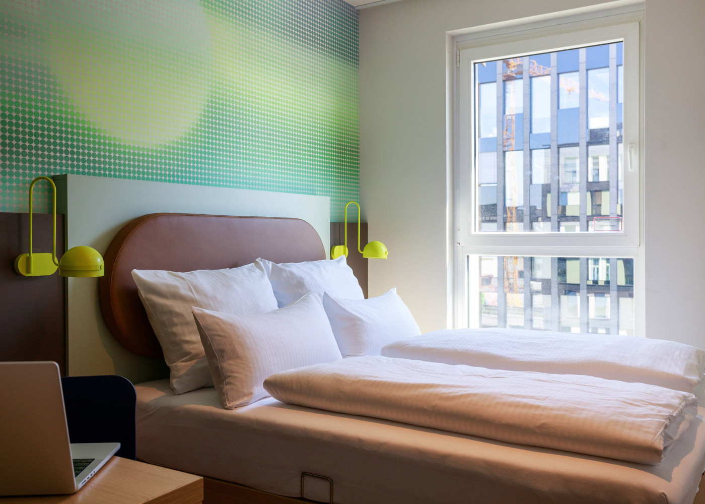 Blick auf ein Hotelzimmer in Frankfurt mit einem Bett vor einem großen Fenster und einem Schreibtisch mit Laptop davor.