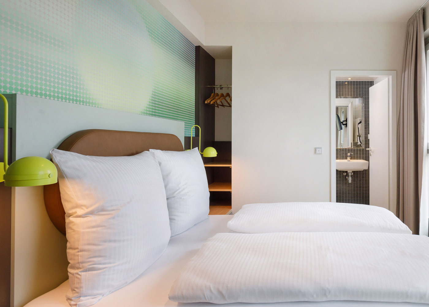 Hotelzimmer mit großem Doppelbett, grüner Wandgestaltung und Blick ins angrenzende Badezimmer mit Spiegel.